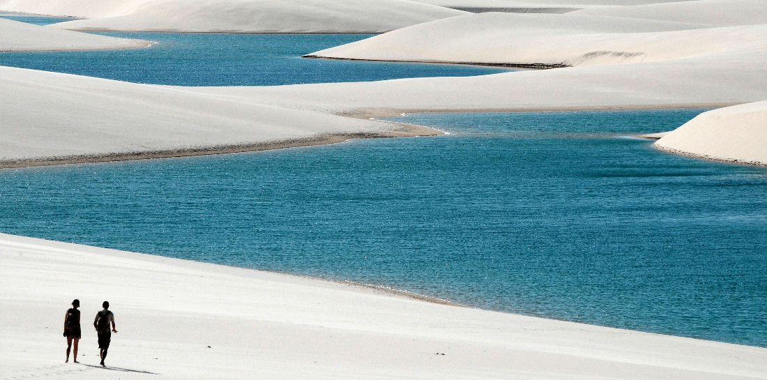 The image shows two people walking along the edge of a body of vibrant blue water, surrounded by smooth white sand dunes that create a striking contrast. The scene is serene and somewhat surreal, reminiscent of a desert oasis. The sky is not visible, focusing the view on the interaction between the earthy tones of the sand and the refreshing blue of the water, with the human figures adding a sense of scale and life to the landscape.